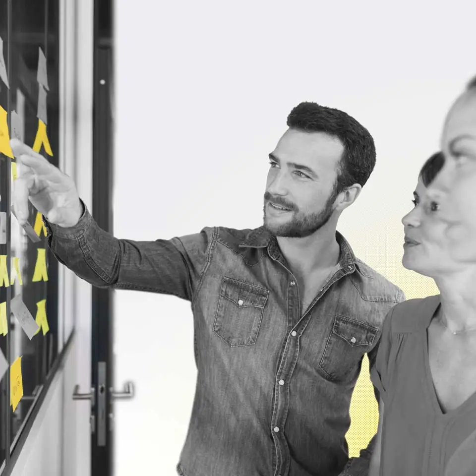A  man and woman looking at sticky notes on a glass wall in a sales workshop